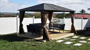 A gazebo with a sloped roof and curtains, surrounded by a white fence, with outdoor furniture and a view of a city skyline. - Regal Residency (Auckland)