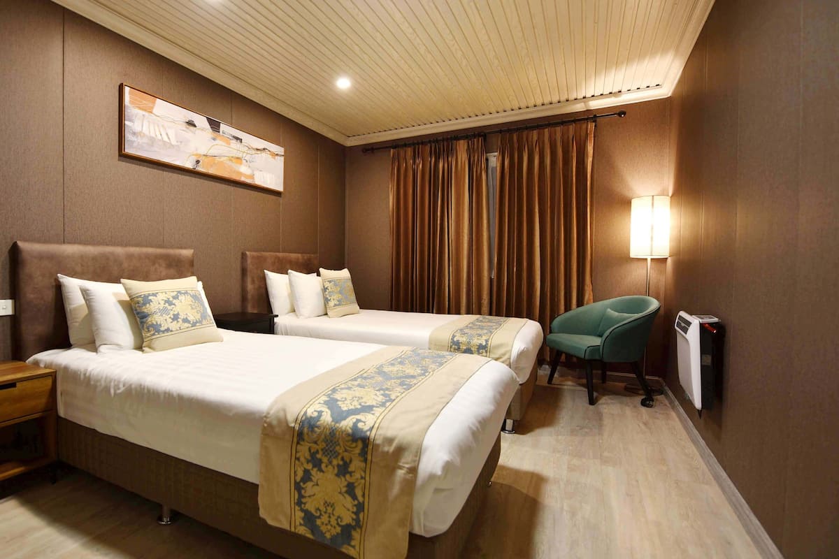 Quarto de hotel com duas camas, uma mesa de cabeceira, uma cadeira, um abajur e um radiador.