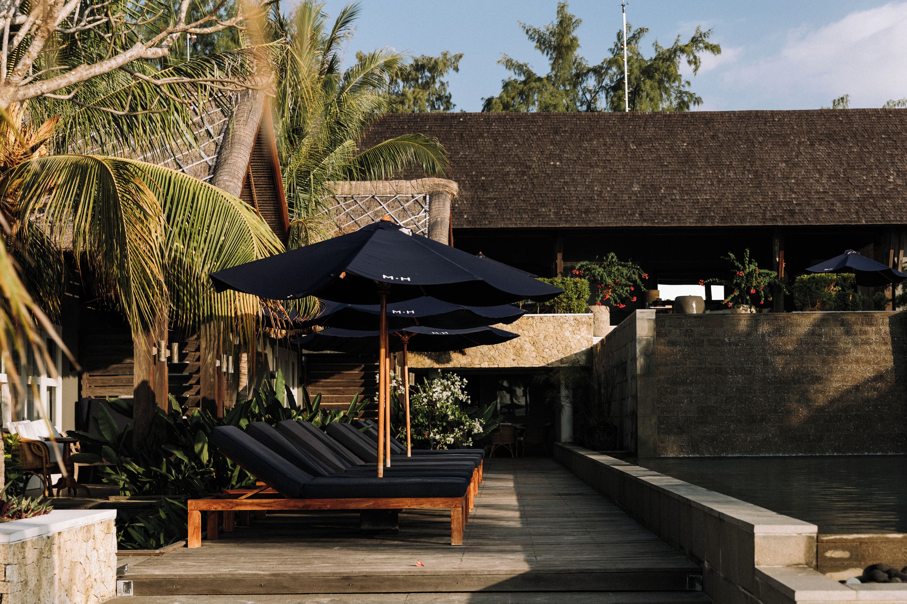 A poolside area with lounge chairs and umbrellas, surrounded by tropical plants and a stone wall.