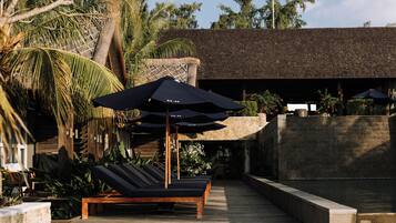 A poolside area with lounge chairs and umbrellas, surrounded by tropical plants and a stone wall.