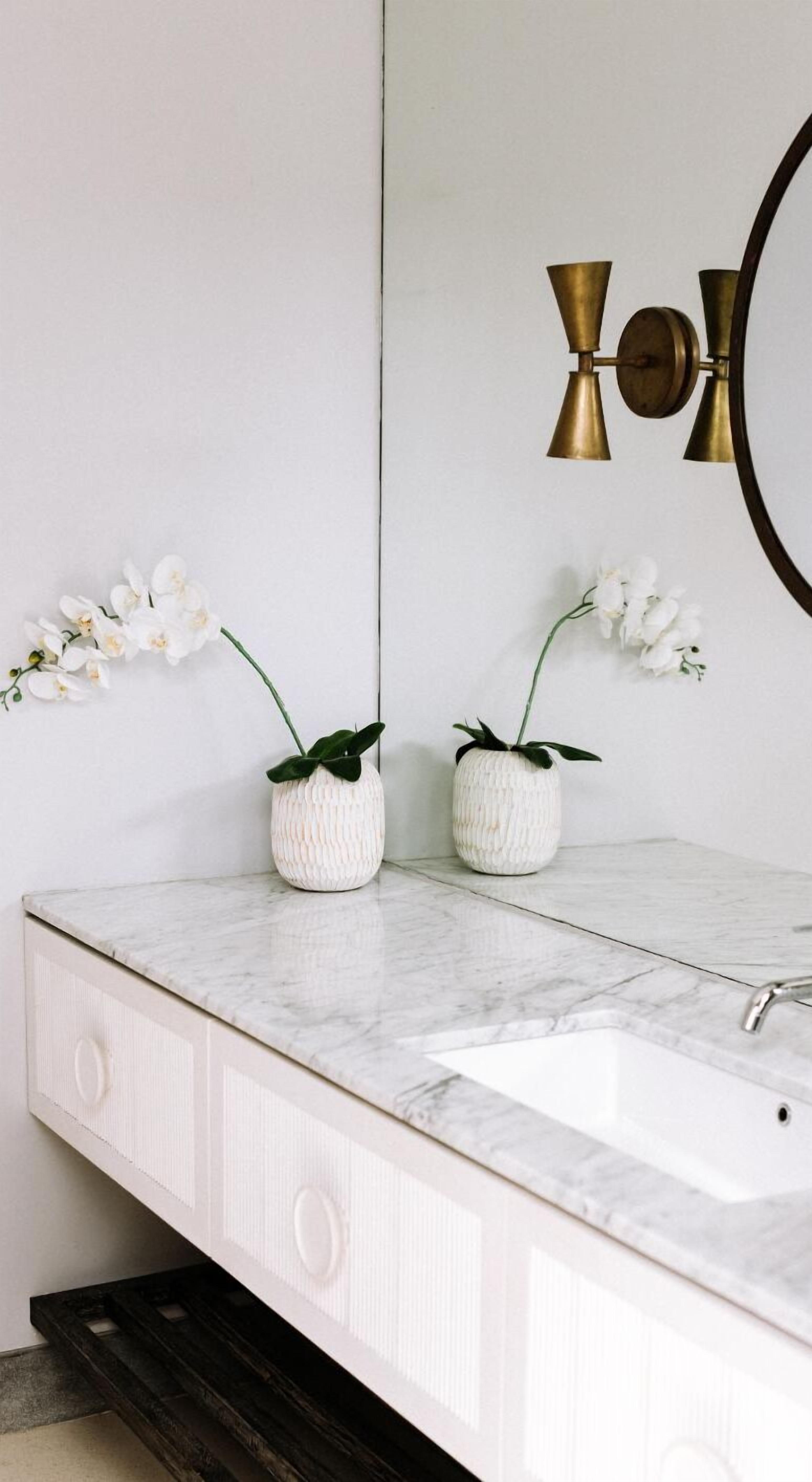 Een badkamer met een wit marmeren wastafel, twee witte keramieken vazen met orchideeën, en een grote spiegel met een ronde spiegel in gouden lijst.