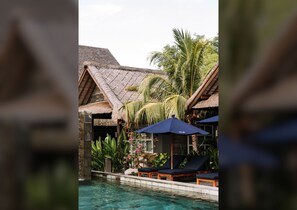 An infinity pool with lounge chairs and umbrellas, surrounded by lush greenery and a thatched-roof structure.