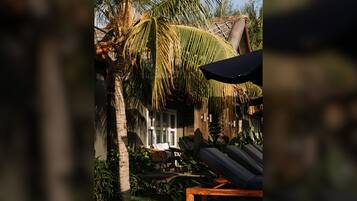 A tropical resort setting with a wooden lounge chair, a thatched-roof structure, and lush greenery.