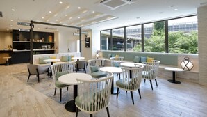 Een moderne binnenruimte met ronde tafels en stoelen, een receptie met een 'Welcome'-bord en grote ramen met uitzicht op groen buiten.
