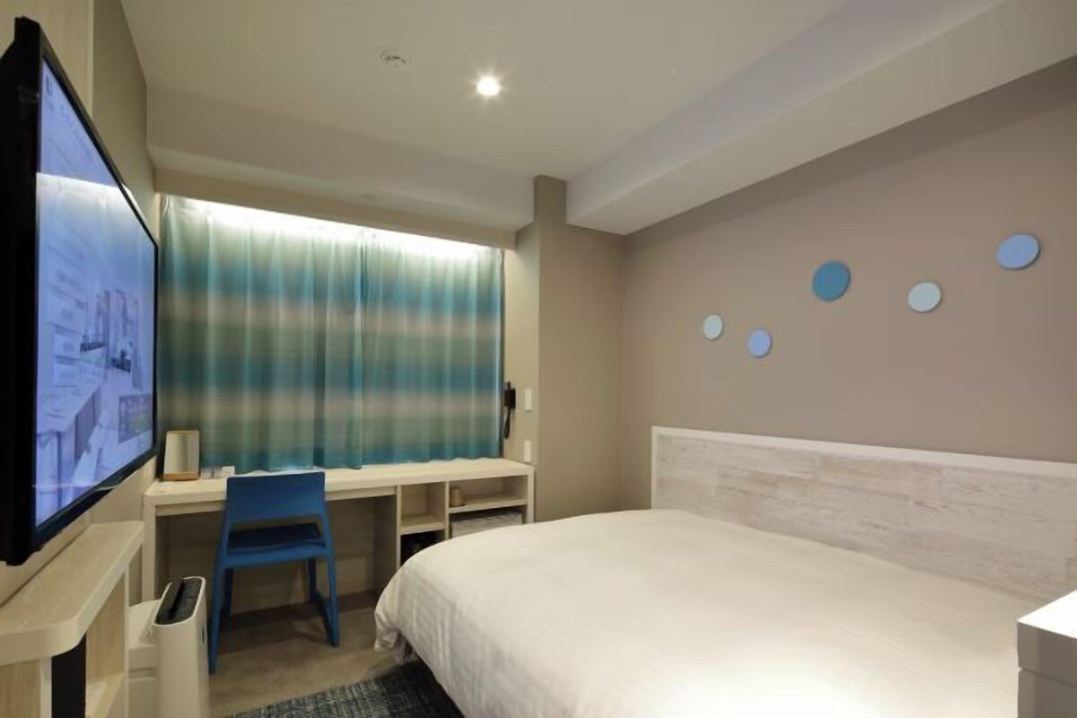 A hotel room with a bed, a desk, a chair, a TV, and a window with curtains.