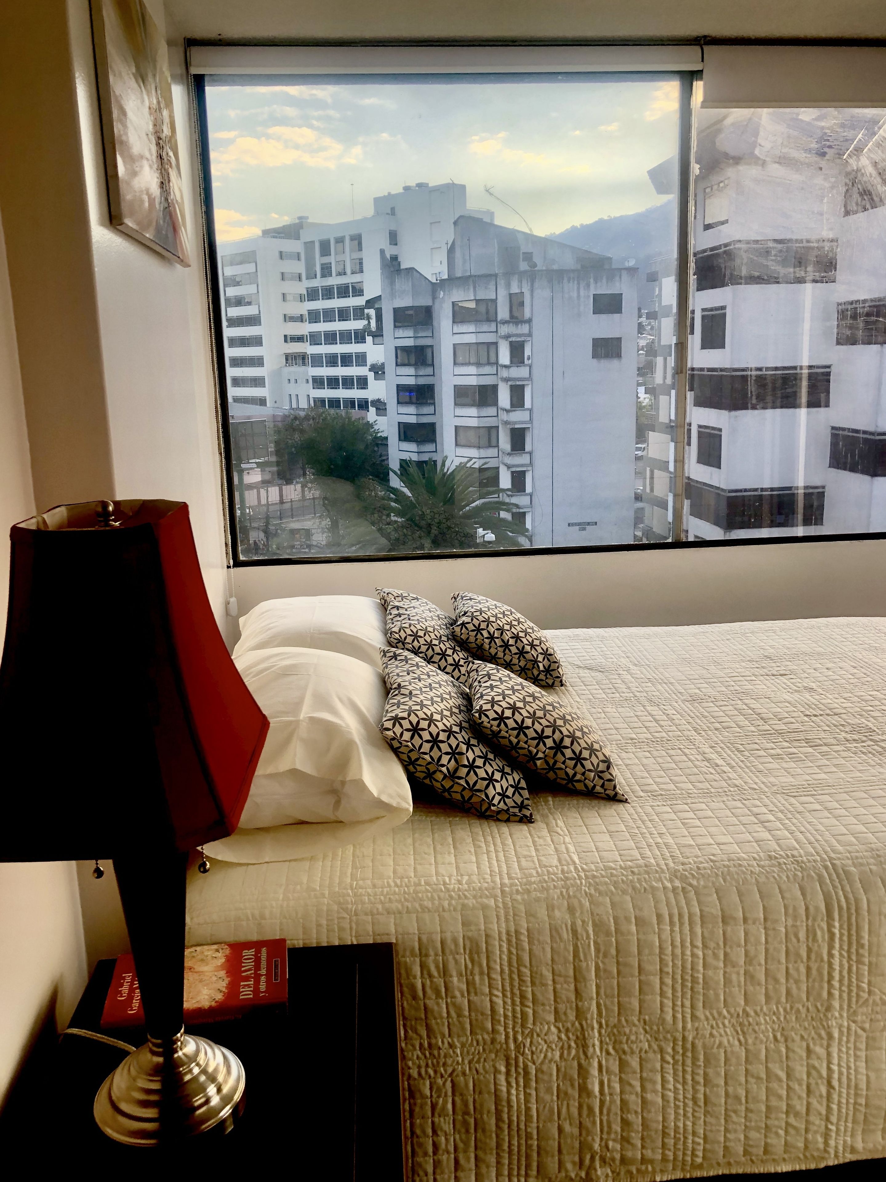 Um quarto com cama, janela com vista para a cidade, abajur e um livro num criado-mudo.