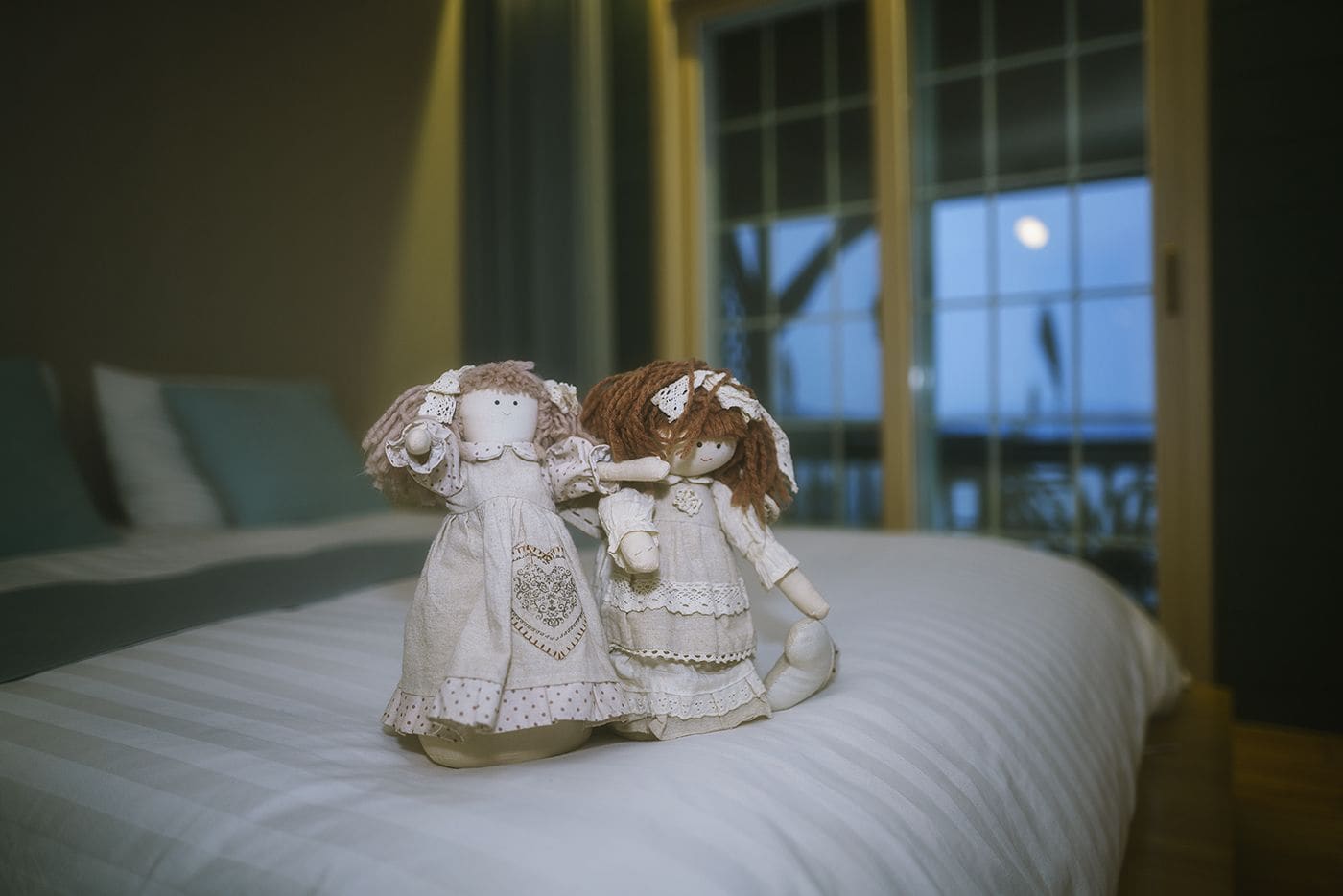 Two dolls on a bed in a room with a large window and a view of the sky.