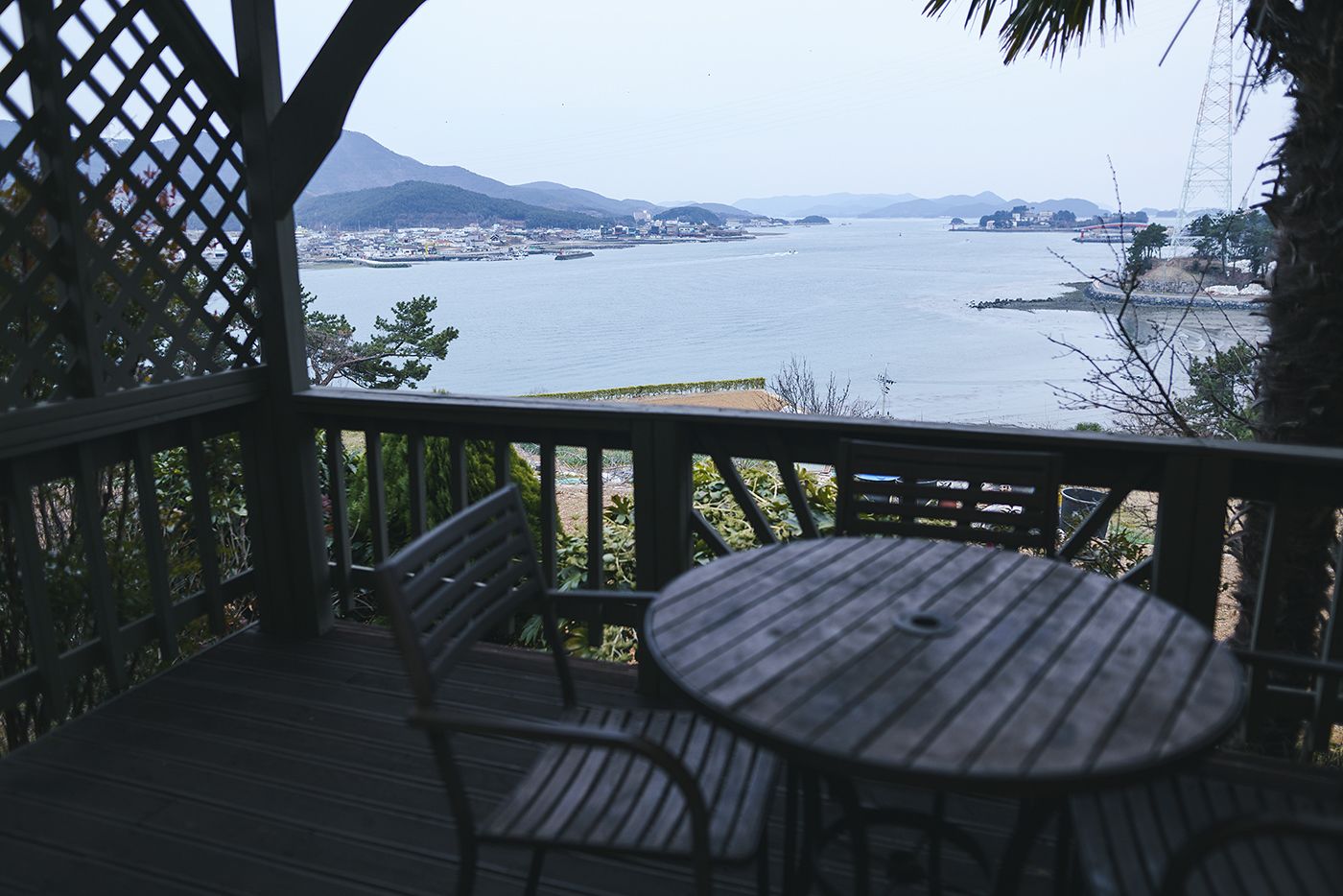 A wooden deck with a round table and two chairs, overlooking a large body of water with distant land and mountains.