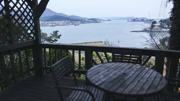A wooden deck with a round table and two chairs, overlooking a large body of water with distant land and mountains.