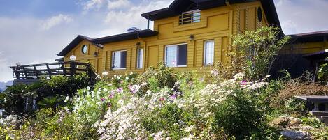 A yellow house with a balcony, surrounded by a garden with various flowers.