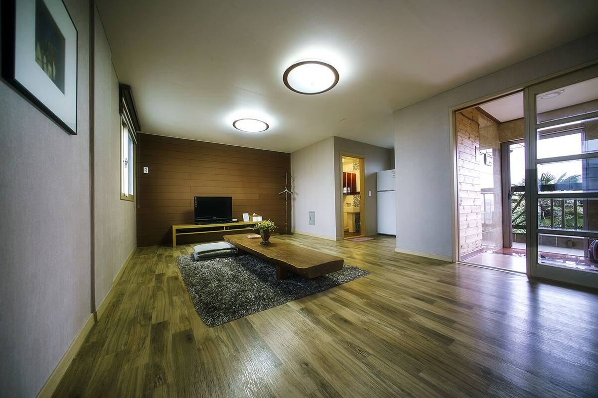 A modern living room with a wooden floor, a flat-screen TV, a dining table, and a kitchen visible through an open door.