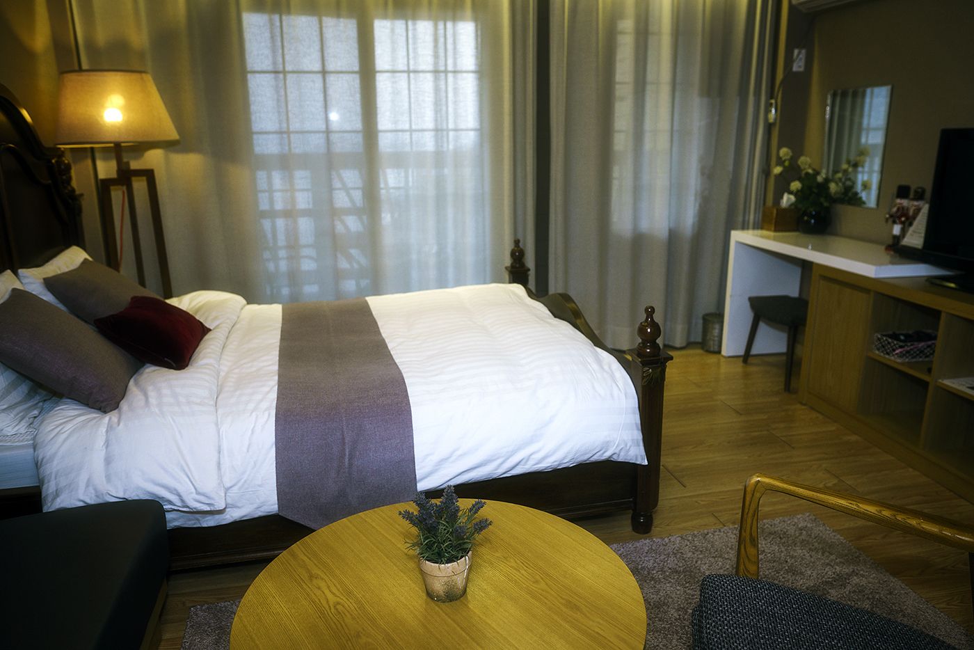 Hotellihuone, jossa on suuri sänky, työpöytä, tuoli, lamppu ja pieni kasvi pyöreällä pöydällä.
