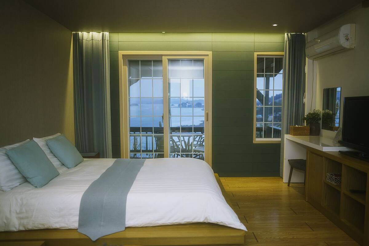 A bedroom with a bed, a desk with a TV, and a view of the outdoors through a window.