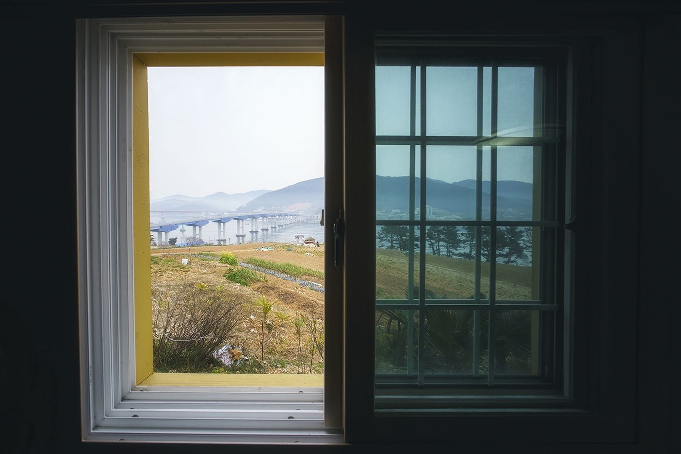 A window with a view of a bridge, a river, and a hilly landscape.