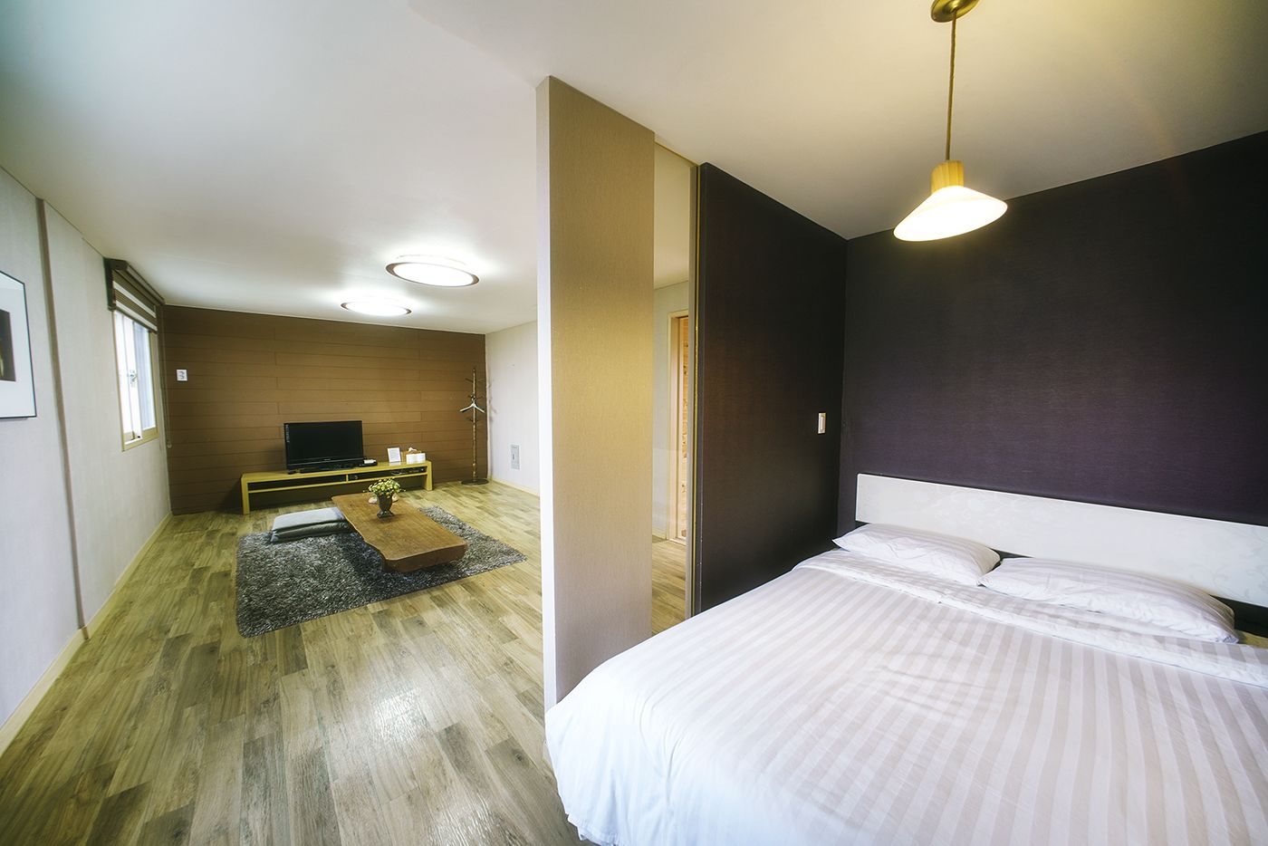 A modern hotel room with a bed, a flat-screen TV, a wooden coffee table, and a dark-colored wall.