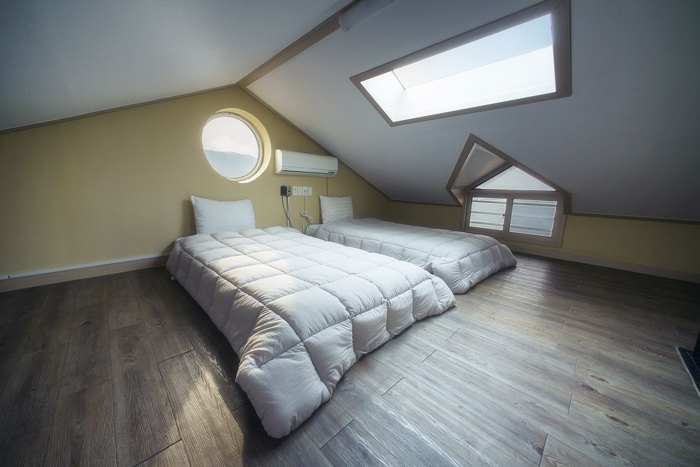 Un dormitorio con techo inclinado, tragaluces, una ventana redonda y una cama con dos colchones blancos.