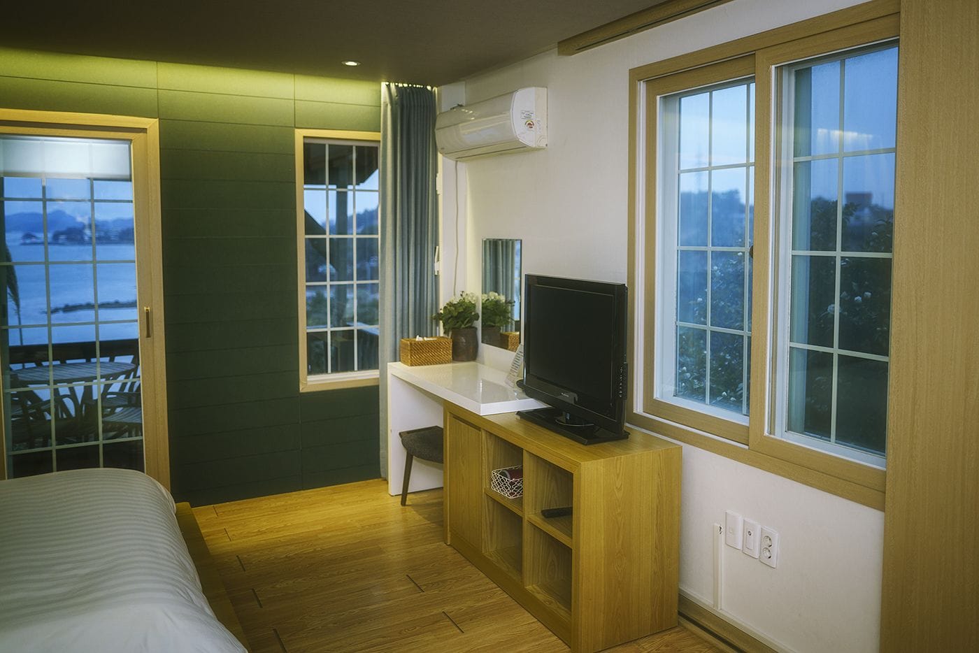 A bedroom with a bed, a desk with a TV, and large windows.