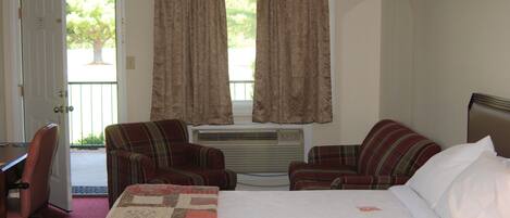 A hotel room with a bed, two plaid chairs, a desk, and a window with curtains.