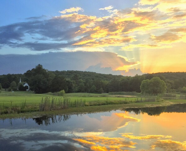 A serene landscape with a body of water reflecting the sky, a church, and a forested hill in the background.
