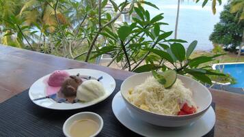 Una mesa con un tazón de comida blanca desmenuzada, un plato de helado y un recipiente pequeño de salsa, frente a un fondo de piscina y palmeras.