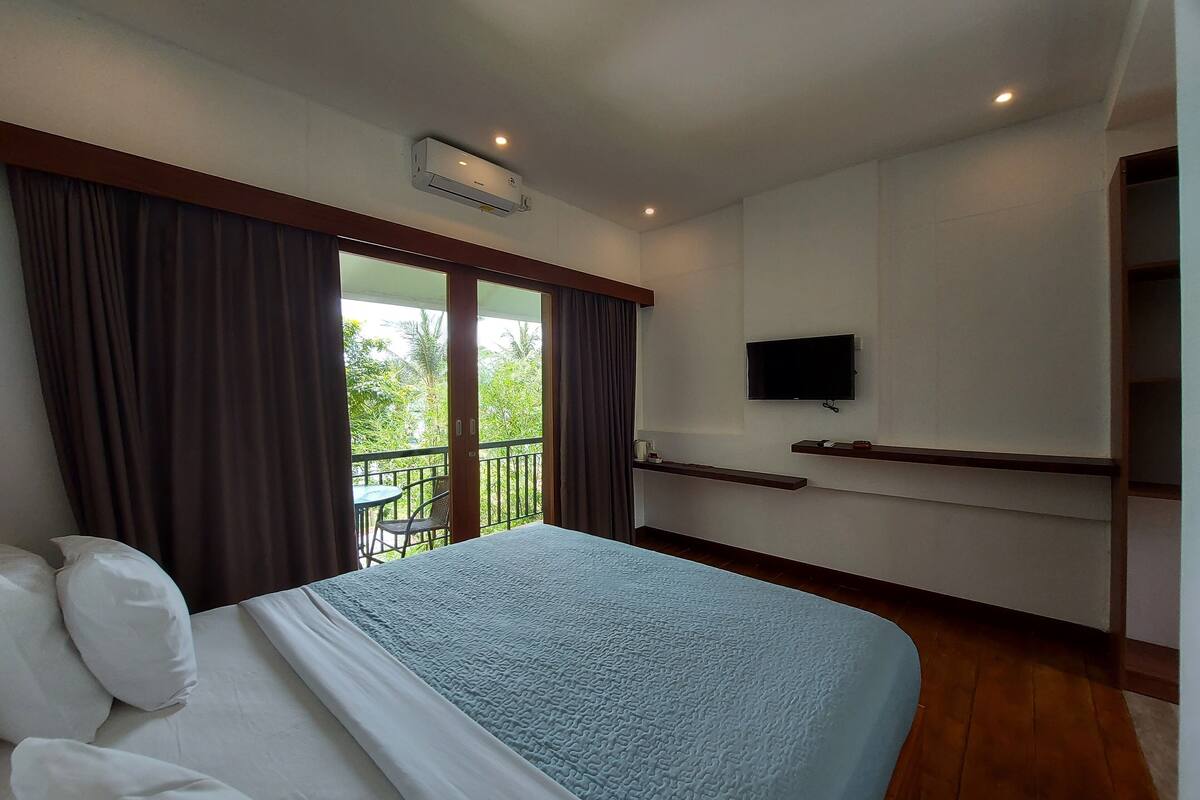 Habitación de hotel con cama, televisor montado en la pared, balcón con cortinas y ventilador de techo.