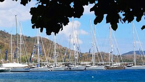 Una marina con diverse barche a vela ormeggiate, circondata da colline e cieli sereni.
