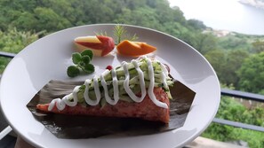 A plate with a wrapped food item, garnished with green leaves and white sauce, set against a scenic outdoor background. - Day Spring (Nangan)