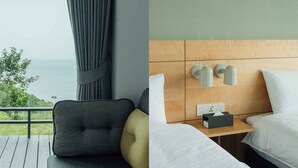 Una habitación de hotel con vista al mar, cabecera de madera, mesita de noche con lámpara y cama con ropa blanca.