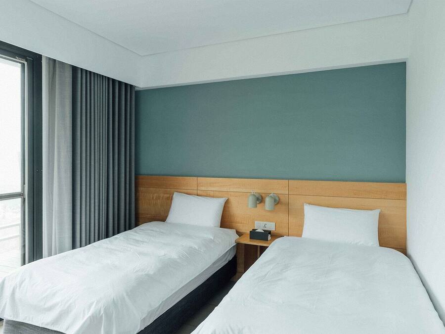 A hotel room with two single beds, a wooden headboard, a nightstand with a lamp, and a window with curtains.