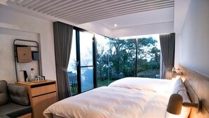 A modern bedroom with a large bed, a bedside table, a chair, and a view of trees through a large window. - Day Spring (Nangan)