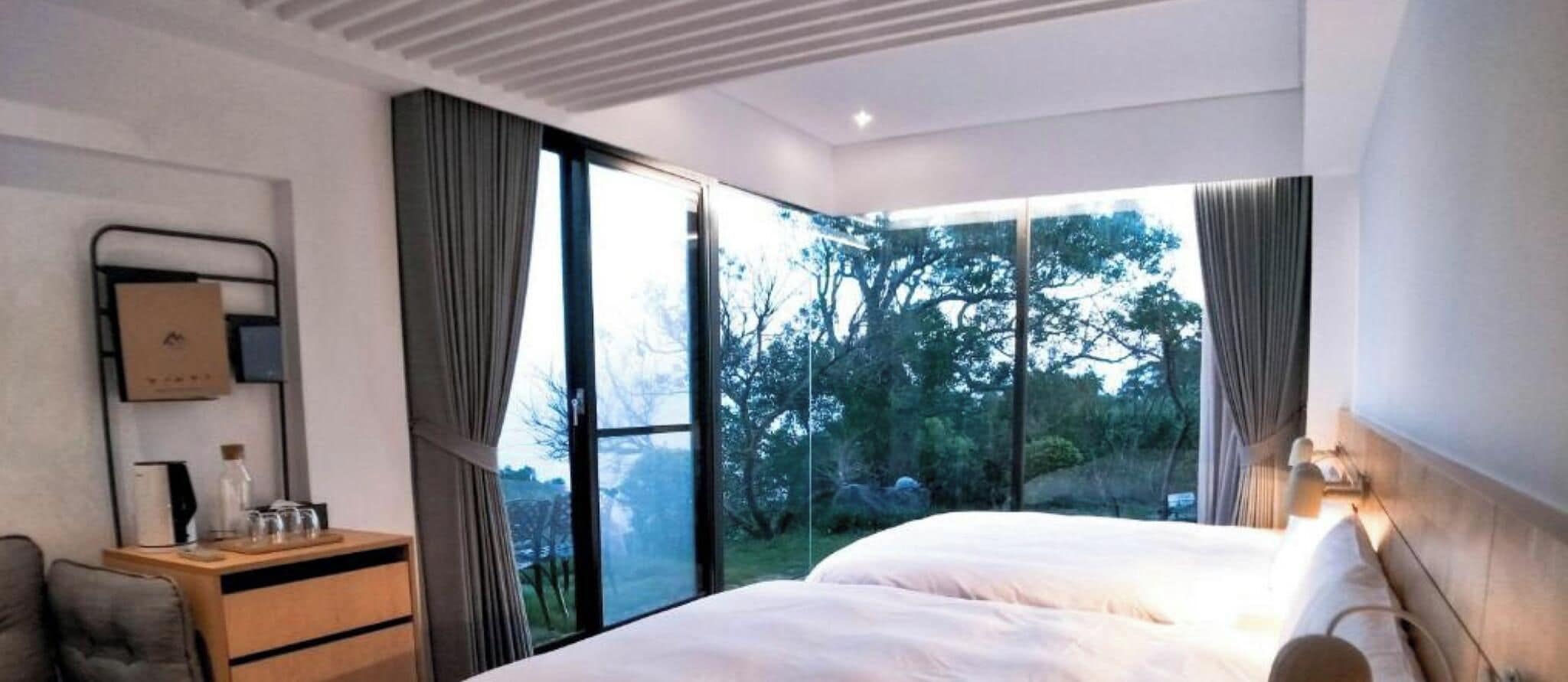 A hotel room with a bed, a desk, a chair, and a large window offering a view of trees.