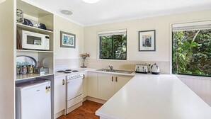 A compact kitchen with white cabinets, a microwave, a stove, and a sink. There are framed pictures on the walls and a window with a view of greenery. - The Cottages (Tamborine Mountain)
