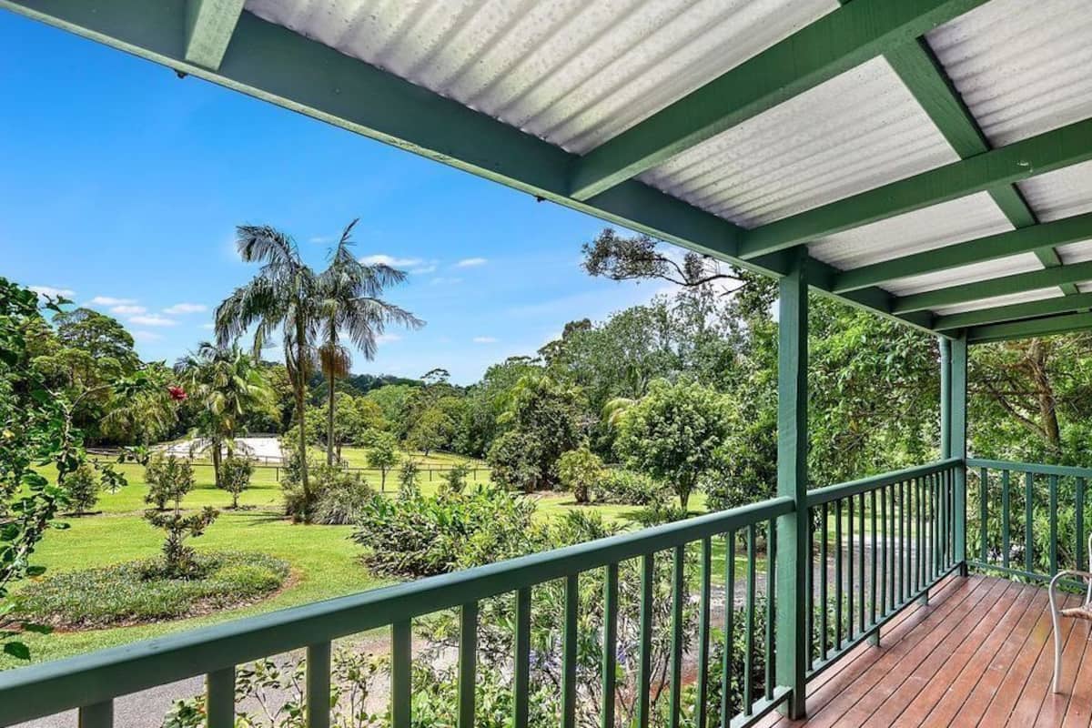 A covered outdoor area with a wooden deck, green railings, and a view of a lush garden with various plants and trees.