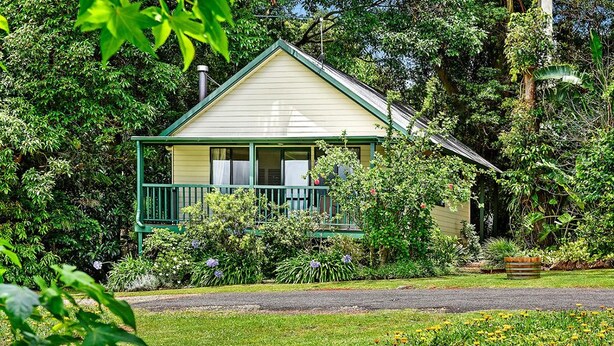 A small, single-story house with a green porch and a white exterior, surrounded by lush greenery and a well-maintained garden. - The Cottages (Tamborine Mountain)