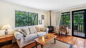 A cozy living room with a sofa, a wooden coffee table, a lamp, a chair, and a large window with blinds. - The Cottages (Tamborine Mountain)
