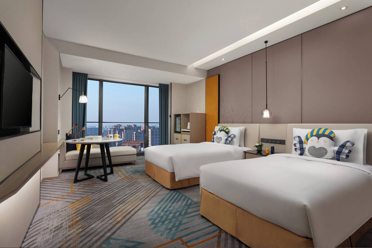 A hotel room with two beds, a desk, and a large window offering a city view.