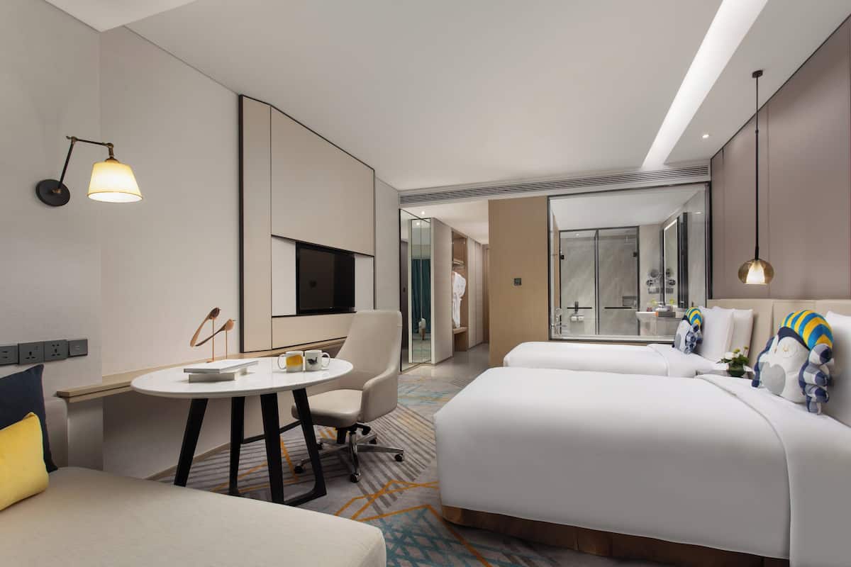 A modern hotel room with a bed, a desk, a chair, and a bathroom visible through an open door.