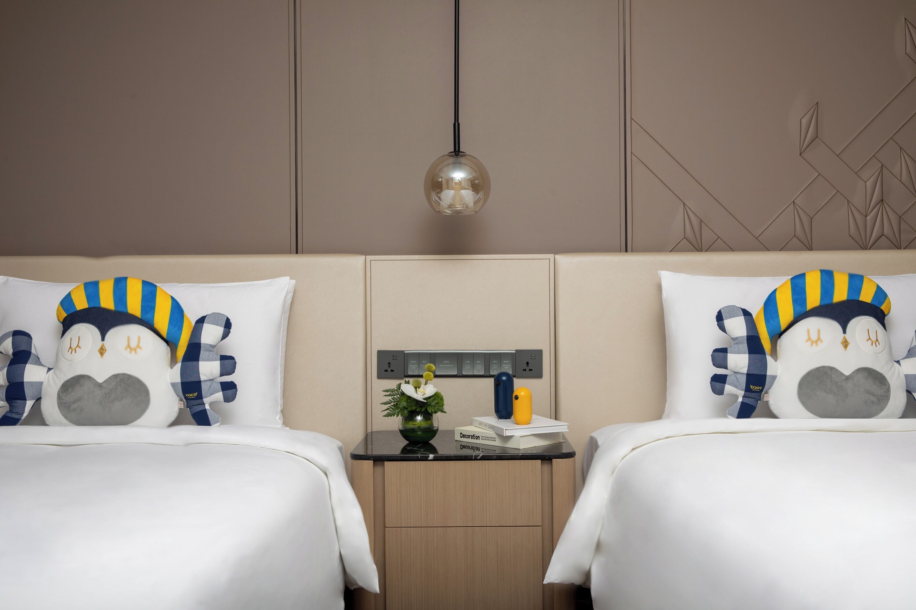 Habitación de hotel con dos camas, cada una con almohadas a juego, una mesita de noche con lámpara y un enchufe en la pared.