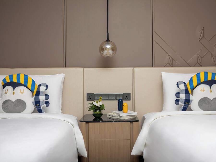 Habitación de hotel con dos camas, cada una con almohadas a juego, una mesita de noche con lámpara y un enchufe en la pared.