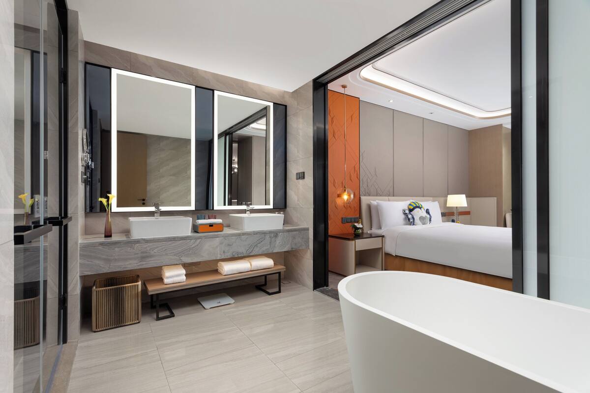 A modern hotel bathroom with a large mirror, a bathtub, a sink, and a bench.