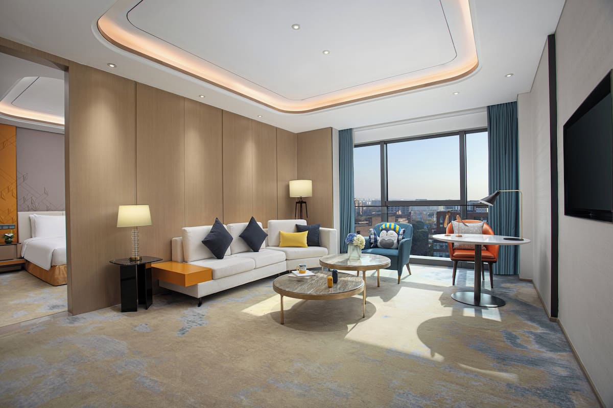 A modern hotel room with a large bed, a sofa, a dining table, and a view of the city.