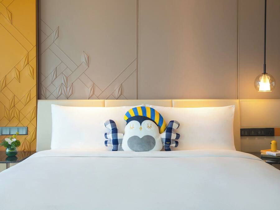 Habitación de hotel con cama, almohada decorativa, mesita de noche, lámpara y pared con diseños geométricos.
