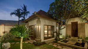 A well-maintained garden with a palm tree, a small pond, and a pathway leading to a house with large windows and a tiled roof. - The Georgina Estate (Nusa Dua)