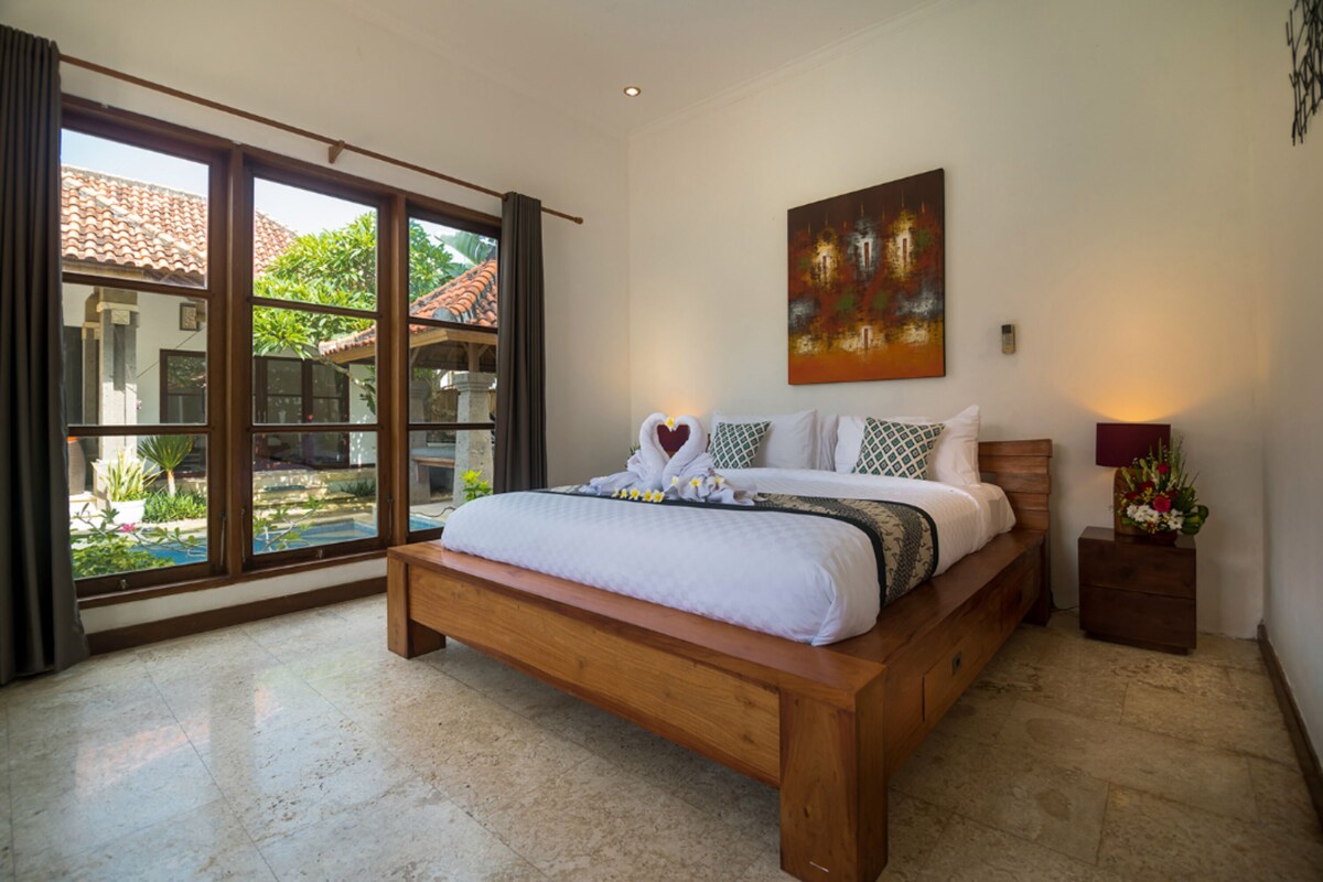A bedroom with a large bed, a painting on the wall, and a view of the outside.