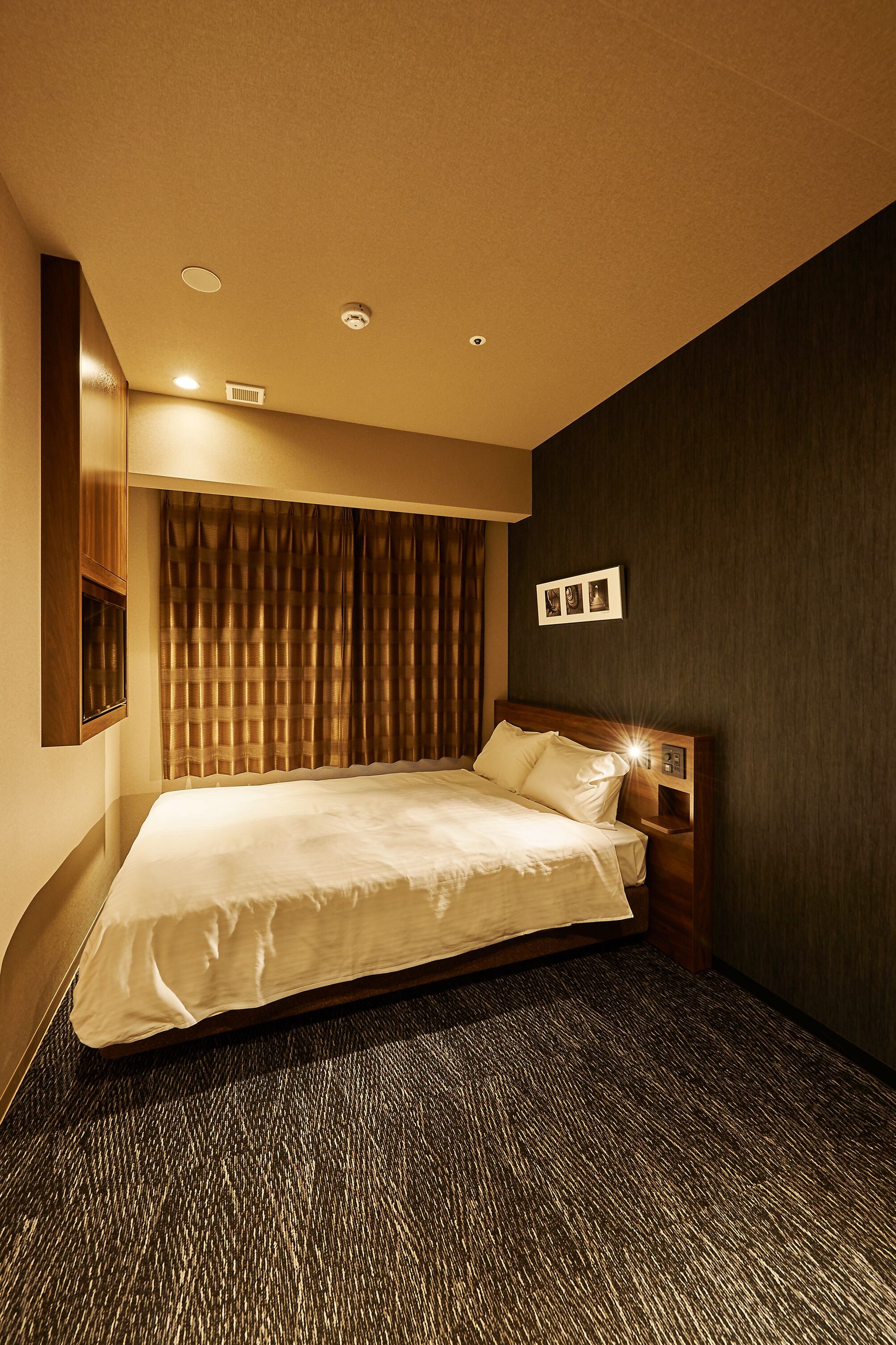 A hotel room with a large bed, a headboard with a light, a wall-mounted TV, and a dark-colored carpet.