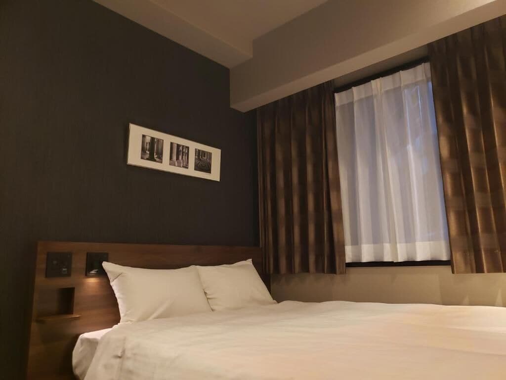 A hotel room with a bed, a headboard, a nightstand, a wall-mounted picture, and a window with curtains.