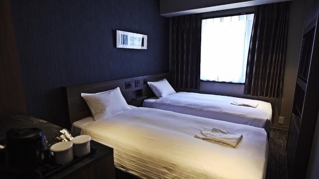 A hotel room with two beds, a desk with a lamp, and a window with curtains.