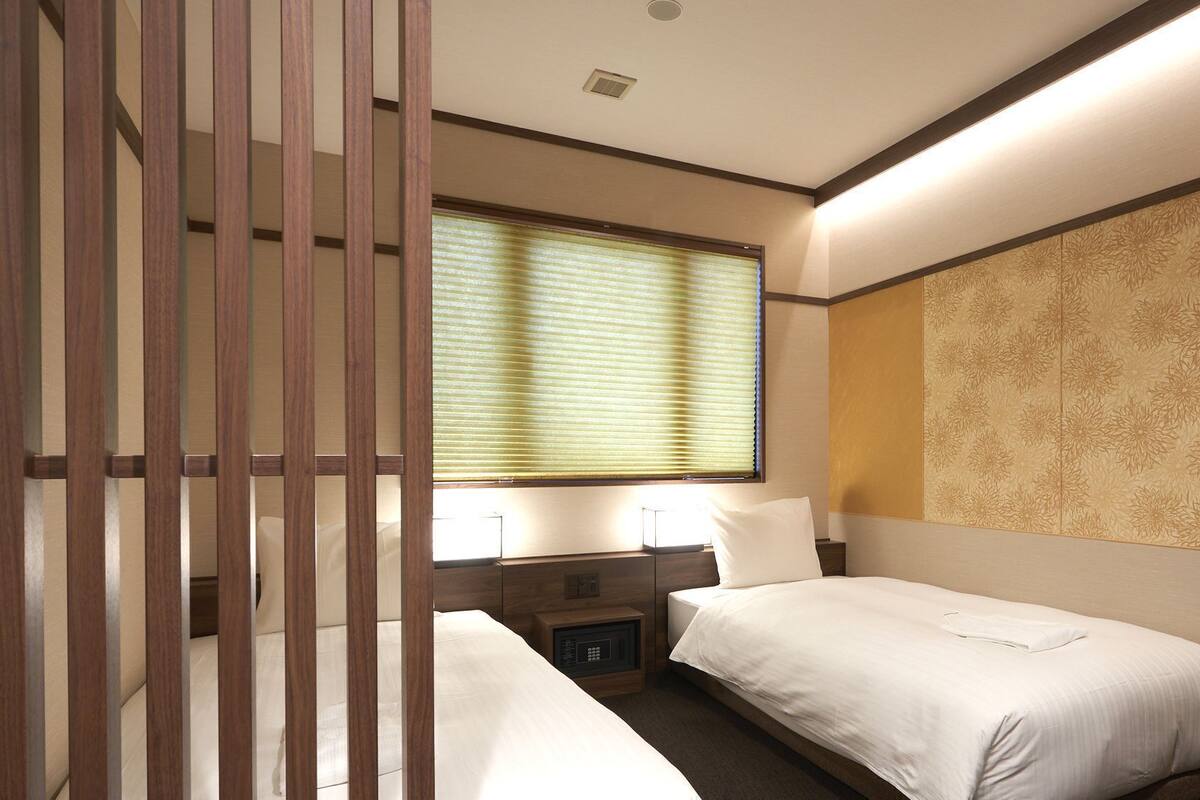Ein Hotelzimmer mit zwei Betten, einem Fenster mit Jalousien, einem Nachttisch mit Lampe und einer dekorativen Wandverkleidung.
