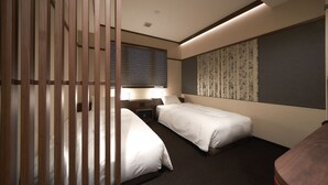 Ett hotellrum med två separata sängar, ett skrivbord med en stol, ett fönster med persienner och en vägg med tapet med ett mönster.