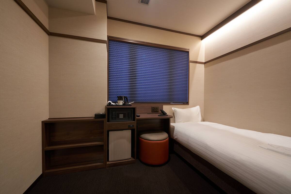 Ein kompaktes Hotelzimmer mit einem Einzelbett, einem kleinen Schreibtisch mit Telefon, einer Mikrowelle und einem Stauraumschrank.